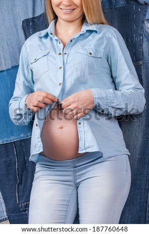 Staying trendy. Cropped image of pregnant woman dressing up her jeans shirt while standing against jeans background