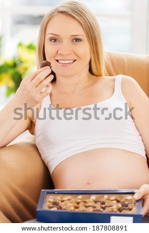 Eating chocolate candies. Beautiful pregnant woman sitting on the chair and eating chocolate candies