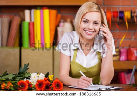 Beautiful florist at work. Beautiful young female florist writing something at note pad and taking on the phone while standing at her working place