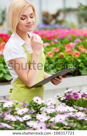 Working with flowers. Beautiful blond hair woman in uniform holding clipboard and looking at the flowers while standing in greenhouse
