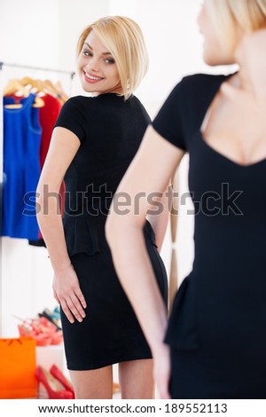 This dress is perfect! Beautiful young blond hair woman in black dress looking at the mirror and smiling