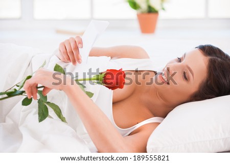 Message from boyfriend. Side view of beautiful young woman lying in bed with red rose and reading letter