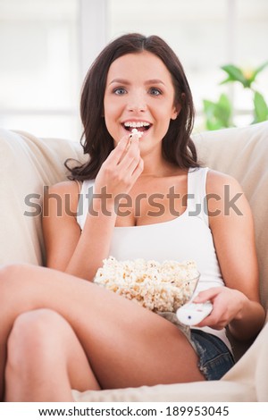 Eating popcorn. Beautiful young woman eating popcorn and watching movie while sitting on sofa