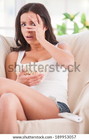 Watching a scary movie. Shocked young woman covering face with hands and watching movie while sitting on sofa