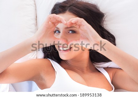 Heart shape. Cheerful young woman lying in bed and stretching and making heart with hands
