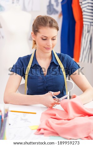 Confident tailor at work. Beautiful young woman with measuring tape on shoulders cutting the cloth with scissors while sitting at her working place