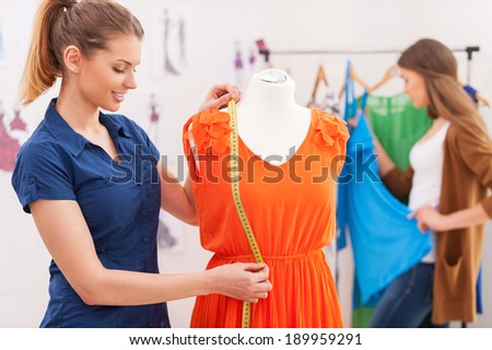 Designer measuring dress. Beautiful female fashion designer measuring dress on the mannequin while her colleague standing on background