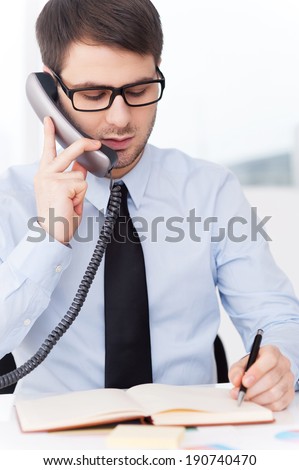 Concentrated on work. Confident young man in shirt and tie talking on the mobile phone and writing something in note pad while sitting at his working place