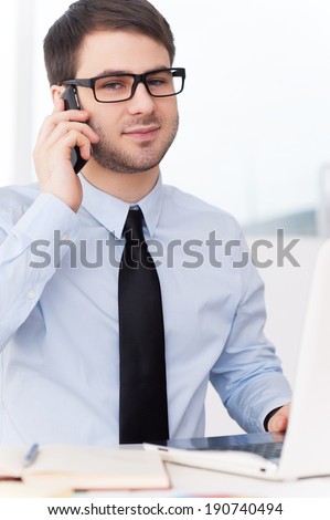 Confident businessman at work. Confident young man in shirt and tie talking on the mobile phone and looking at camera while sitting at his working place