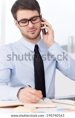 Busy working. Handsome young man in shirt and tie talking on the mobile phone and writing something in note pad while sitting at his working place