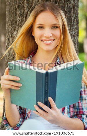 Fresh air and favorite book. Beautiful young woman holding a book and smiling while leaning at the tree in a park