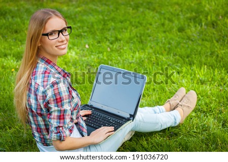 Surfing the net in nature. Beautiful young female student working on laptop and looking over shoulder while sitting on the grass in park