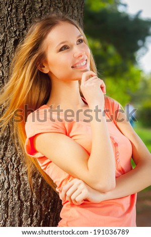 Thoughtful beauty. Side view of beautiful young woman holding hand in hair and smiling while leaning at the tree