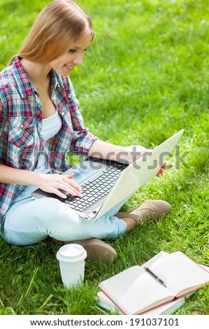 Preparing to exams outdoors. Beautiful young female student working on laptop and smiling while sitting in a park with books around her