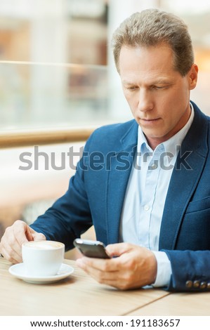 Taking time for coffee break. Confident mature man in formalwear drinking coffee and typing a message on mobile phone while sitting in restaurant