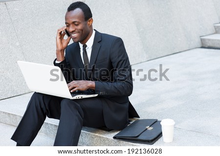 Business is his life. Cheerful young African man in formal wear talking on the mobile phone and working on laptop while sitting on outdoors staircase