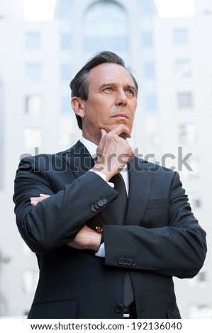 Thinking about solutions. Thoughtful mature man in formal wear holding hand on chin and looking away while standing outdoors