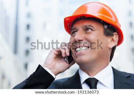 Happy contractor. Cheerful mature man in formal wear and hardhat talking on the mobile phone and smiling while standing outdoors