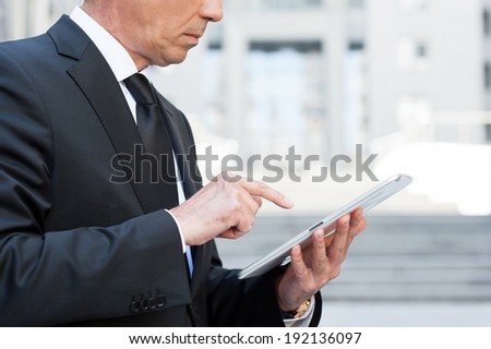 Working on tablet. Side view cropped image of confident senior man in formal wear working on digital tablet while standing outdoors