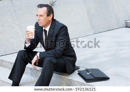 Relaxing after hard working day. Thoughtful mature man in formal wear holding coffee cup and looking away while sitting at the staircase