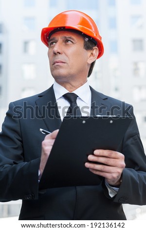 Confident foreman. Thoughtful mature man in formal wear and hardhat writing something in clipboard and looking away while standing outdoors