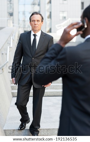 Business people. Rear view of African man in formal wear talking on the mobile phone while another businessman walking by stairs on background