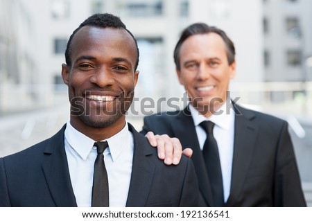 Business support. Confident senior man in formal wear holding hand on shoulder of young African man that looking at camera and smiling