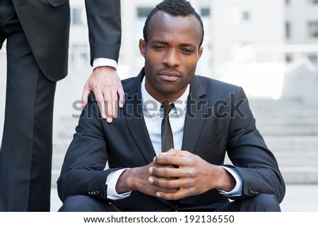 He needs a friendly support. Frustrated young African man in formal wear sitting on staircase while someone touching his shoulder with hand