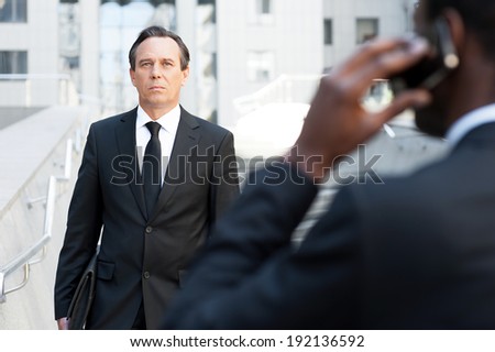 Confident business expert. Rear view of African man in formal wear talking on the mobile phone while another businessman walking by stairs on background