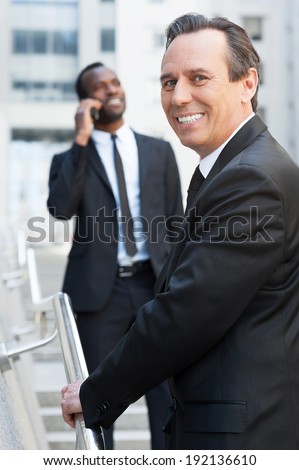 He is the best in his business. Cheerful senior man in formal wear moving up by stairs and looking over shoulder while African man talking on the mobile phone in background