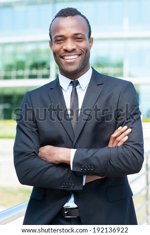 Confident and successful businessman. Handsome young African man in full suit keeping arms crossed and looking at camera while standing outdoors