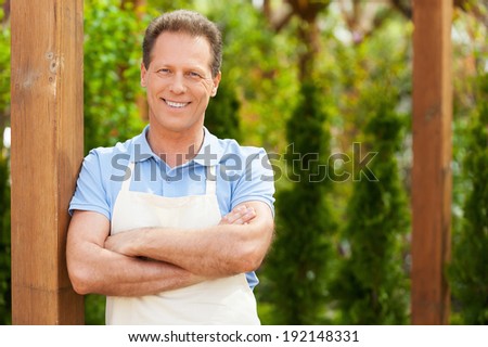 Man in a greenhouse. Handsome mature man in apron keeping arms crossed and looking at camera while standing in a greenhouse