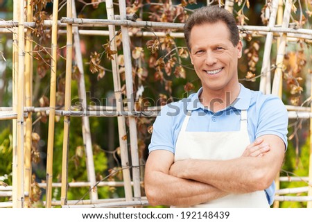 I love to work with plants. Handsome mature man in apron keeping arms crossed and smiling while standing in a greenhouse
