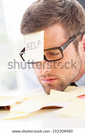Help! Frustrated young man in formal wear with adhesive note on his forehead leaning his head at the table