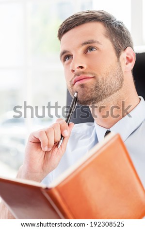 Business dreams. Thoughtful young man in shirt and tie holding note pad and touching chin with pen while sitting at his working place