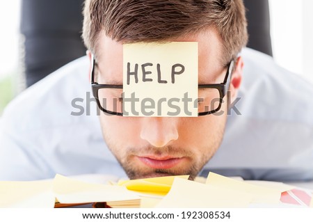 He needs help. Frustrated young man in formal wear with adhesive note on his forehead leaning his head at the table