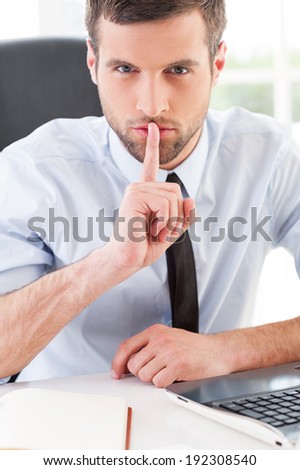 Keep silence! Confident young man in formal wear holding finger on mouth and looking at camera while sitting at his working place