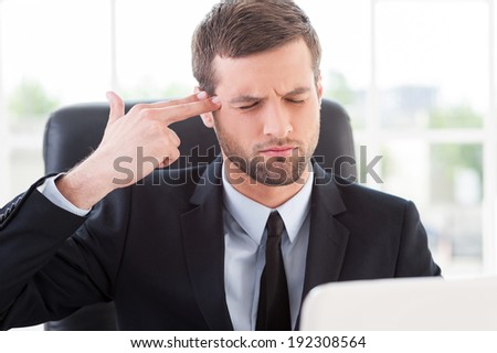 Stressful day. Depressed young man in formal wear touching his temple with finger gun and keeping eyes closed while sitting at his working place