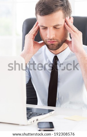 Feeling tired and depressed. Depressed young man in shirt and tie holding head in hands and keeping eyes closed while sitting at his working place