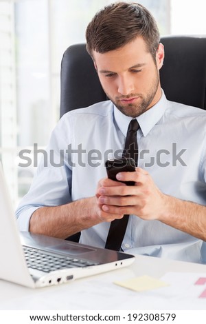 Typing business message. Handsome young man in formal wear looking at his mobile phone while sitting at his working place