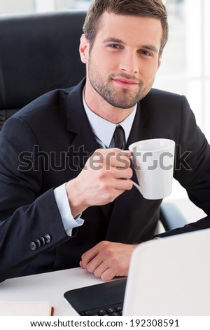 Taking a coffee break. Top view of confident young man in formalwear drinking coffee and smiling while sitting at his working place