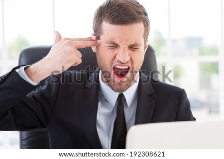 Emotional stress. Furious young man in formal wear touching his temple with finger gun and keeping eyes closed while sitting at his working place
