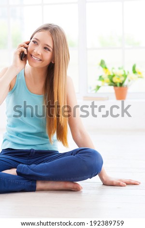I am so glad to hear you! Cheerful teenage girl talking on the mobile phone and smiling while sitting on the floor at her apartment