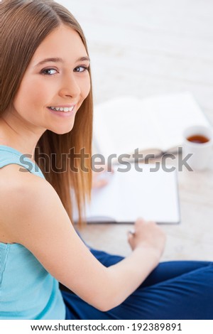 Studying at home. Top view of cheerful teenage girl looking at camera and smiling while books and note pad laying near her
