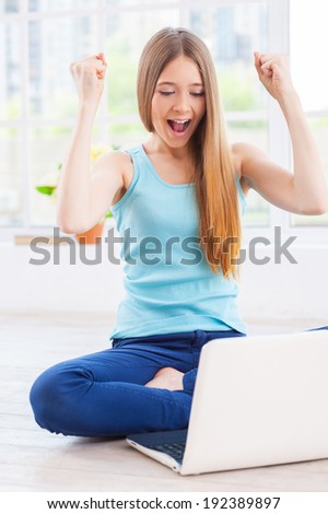 I won! Cheerful teenage girl looking at laptop and keeping arms raised while sitting on the floor at her apartment