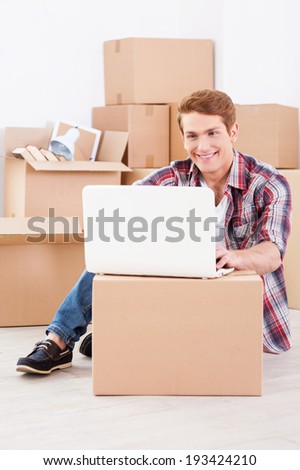 Searching a moving company in the net. Cheerful young man sitting on the floor and working on laptop while cardboard boxes laying in the background