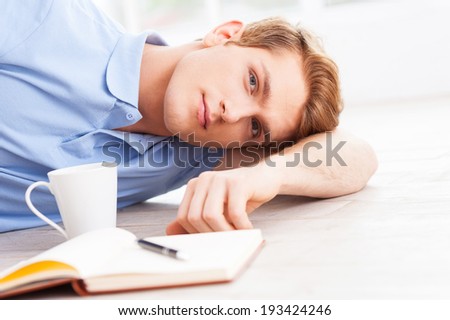 Relaxing at home. Handsome young man lying on the floor and looking at camera with note pad and cup of coffee near him