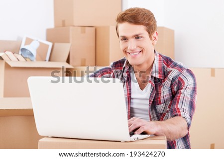 In search of a good moving company. Handsome young man sitting on the floor and working on laptop while cardboard boxes laying in the background