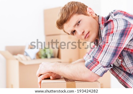 Tired of moving. Side view of depressed young man leaning at the cardboard box and expressing negativity while more boxes laying in the background