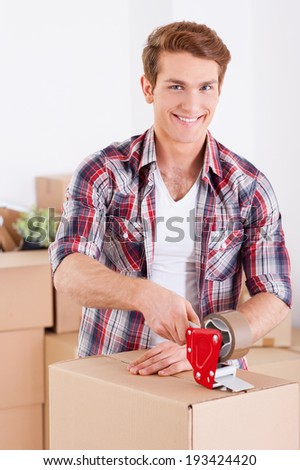 Getting ready for moving. Handsome young man packing boxes and smiling while other cardboard boxes laying in the background